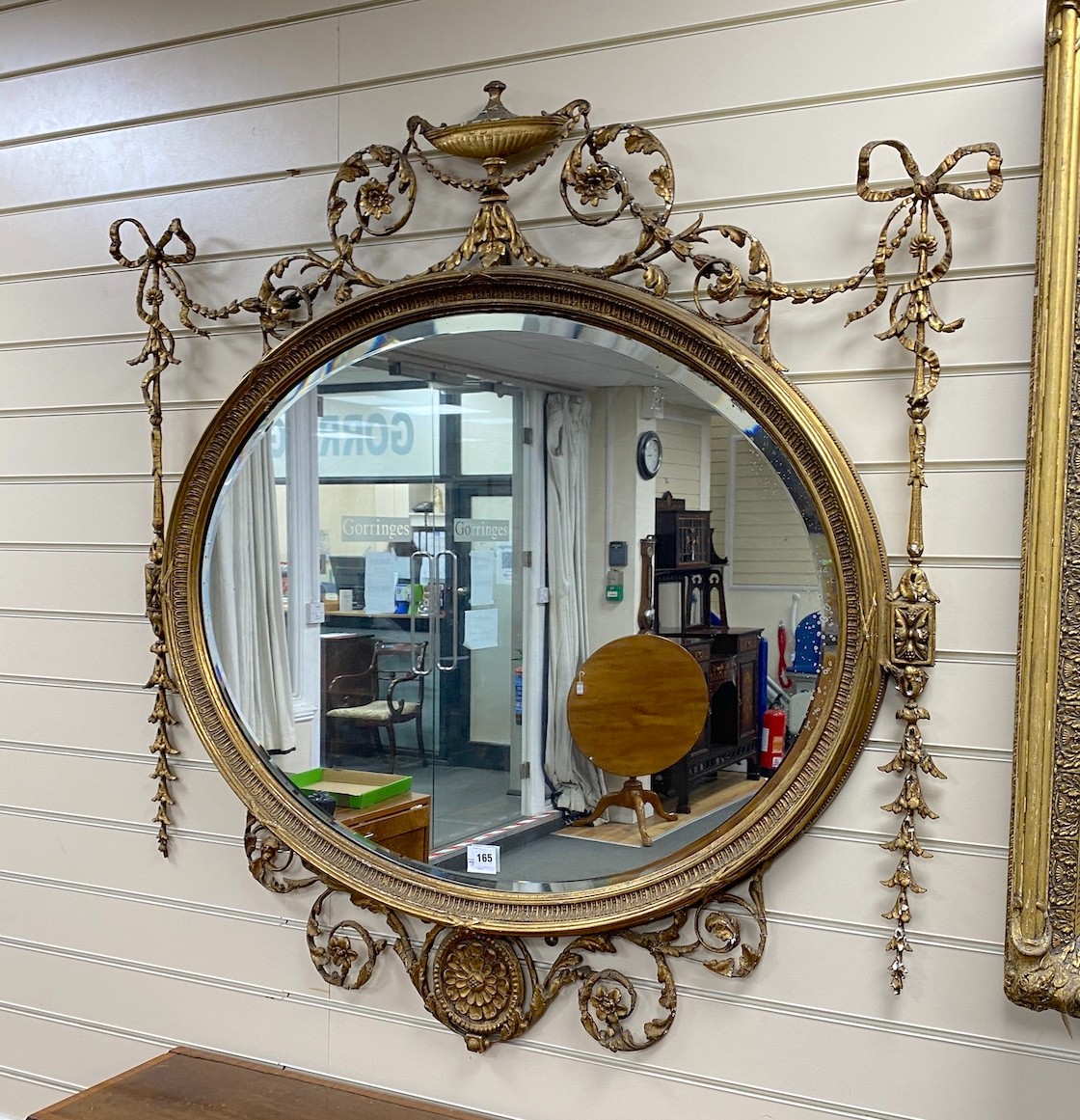 A late Victorian Adam Revival oval wall mirror, with urn, foliate scroll, ribbon and harebell motifs, and bevelled glass plate, width 119cm, height 118cm